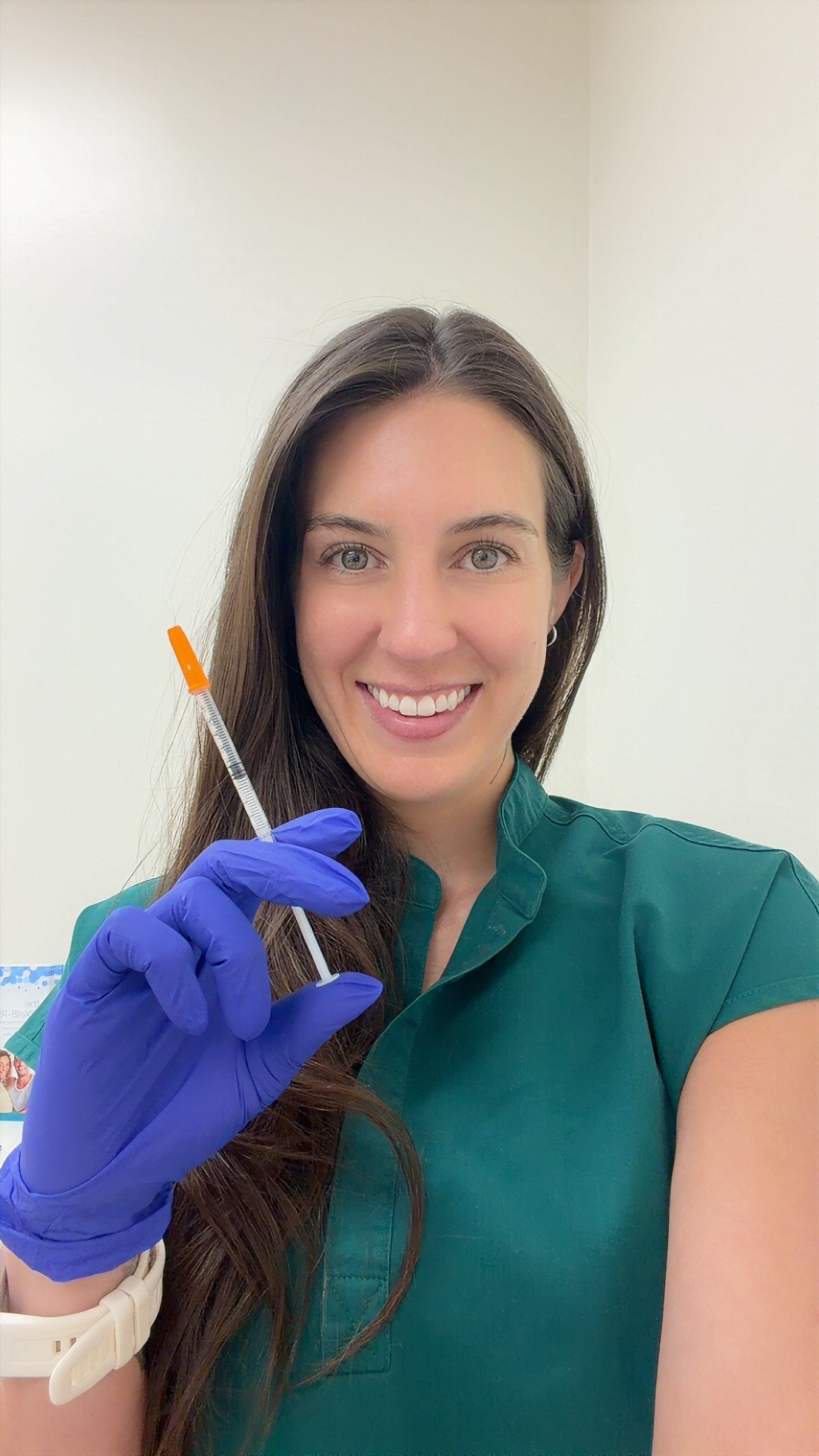 Smiling woman in green scrubs wearing blue gloves holds a medical swab against a neutral background.